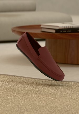 Men’s dark red leather loafer with black sole floating above textured beige rug, with wooden table and stacked books in background.