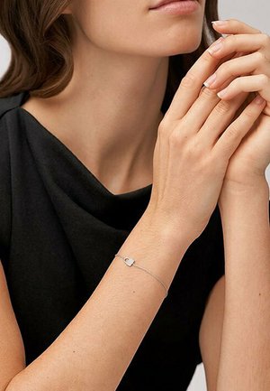 Bracciale d'argento con un piccolo ciondolo a forma di lucchetto; catena delicata, finitura liscia, indossato su una mano con le dita incrociate, che si abbina a un top nero.