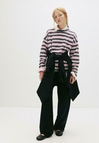 Stribet langærmet skjorte i pink og grå, parret med brede sorte bukser og en bundet sort sweater om taljen. Flade sorte sko.