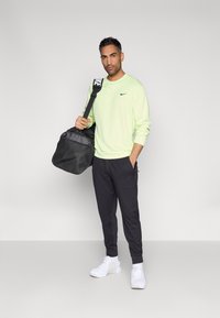 Ανοιχτοπράσινο μακρυμάνικο πουκάμισο με μαύρο λογότυπο Nike, μαύρο ελαστικό παντελόνι με στενή εφαρμογή και λευκά αθλητικά παπούτσια. Κρατάει μια μαύρη τσάντα γυμναστηρίου με ρυθμιζόμενο ιμάντα.