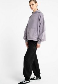 Oversized fioletowa bluza z kapturem z przednią kieszenią, zestawiona z czarnymi dresami. Obie części charakteryzują się luźnym krojem i minimalnymi detalami logo.