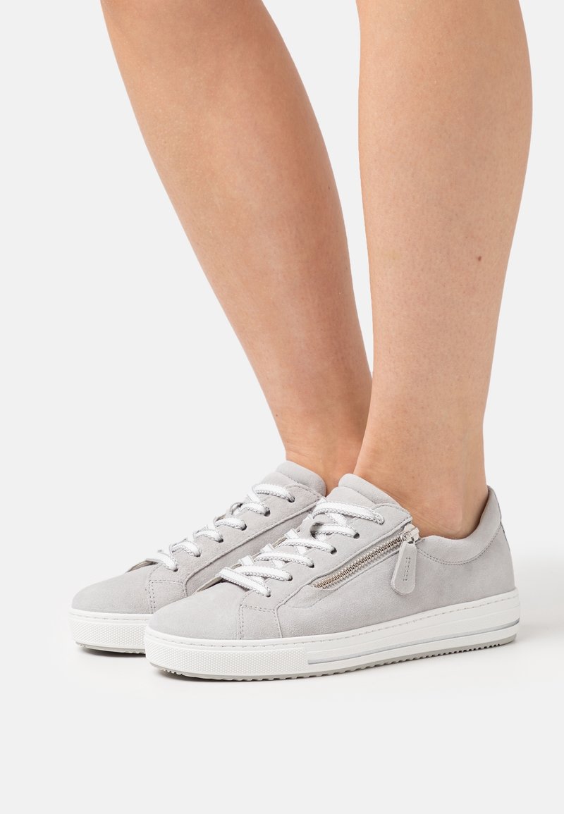 Personne portant des baskets en daim gris clair avec des lacets blancs et une fermeture éclair latérale, debout devant un fond blanc uni.