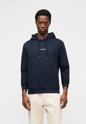 Bărbat purtând un hanorac albastru închis cu textul "LES DEUX", asortat cu pantaloni bej deschis, stând într-un decor simplu, gri deschis.