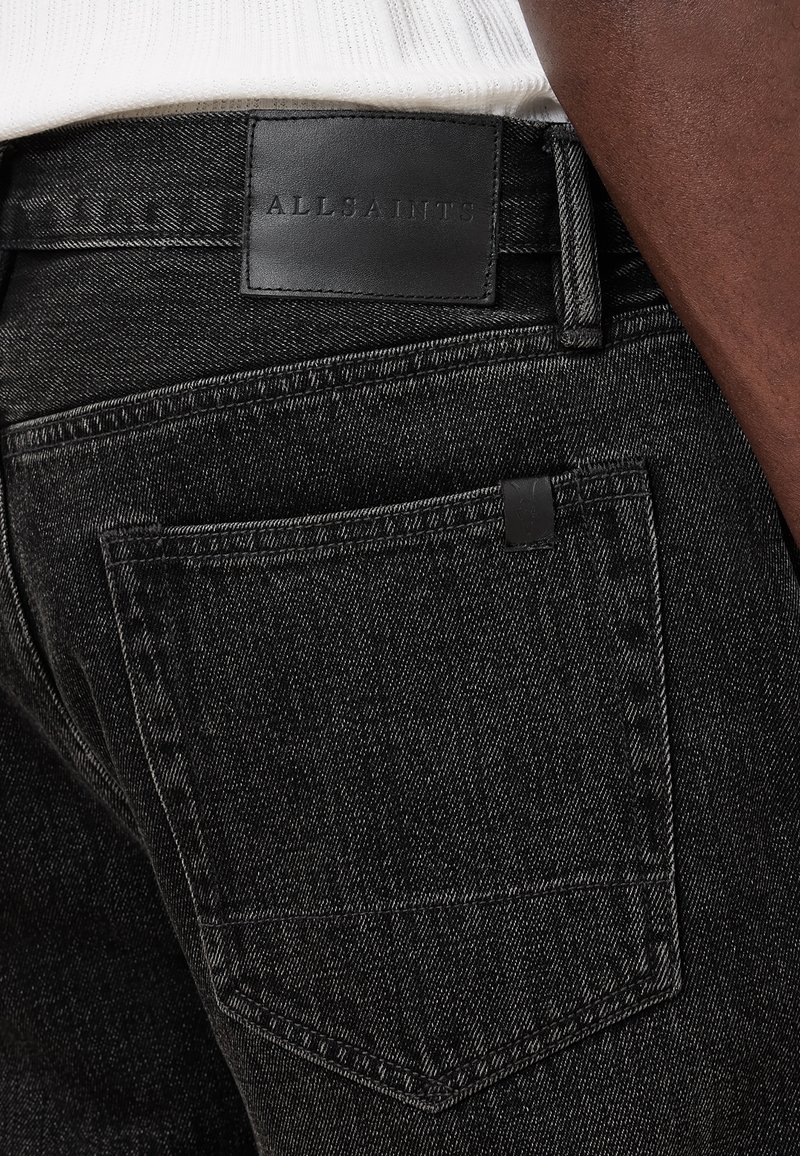 Jean en denim noir à texture lisse, avec une coupe slim, une poche arrière et un patch logo en cuir noir à la taille.