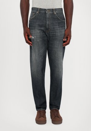 Hombre que lleva jeans negros desgastados con ligeras desgastes en el muslo izquierdo, combinados con zapatillas marrones, de pie contra un fondo blanco liso.