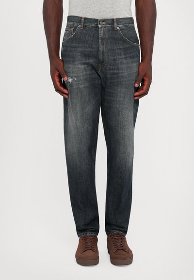 Man wearing faded black jeans with slight distressing on left thigh, paired with brown sneakers, standing against plain white background.
