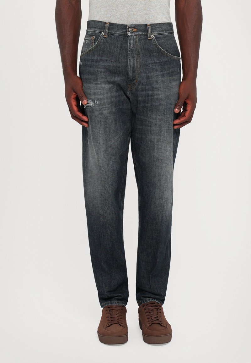 Uomo che indossa jeans neri sbiaditi con lievi strappi sulla coscia sinistra, abbinati a sneakers marroni, in piedi su uno sfondo bianco semplice.