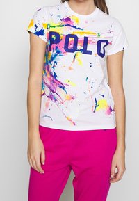 Bílá tričko s barevnými skvrnami v růžové, modré, žluté a fialové, s výrazným námořnickým textem "POLD" přes prsa.