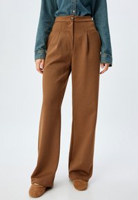 Pantalons taille haute à chevrons marron avec des plis, dotés d'une fermeture à bouton. Associés à une chemise en denim et des mocassins marron.