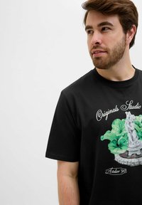 Camiseta negra de manga corta con un estampado gráfico de una estatua rodeada de hojas verdes y el texto "Originals Studio Atelier 90."