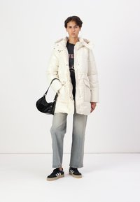 Jachetă puffer albă cu glugă, design matlasat și nasturi pe buton. Asortată cu un tricou grafic negru, blugi ușor albaștri și adidași negri.