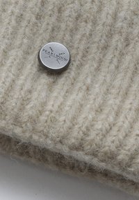 Šviesiai smėlinis vilnos skrybėle, su raštuotu tekstūru. Prisegtas sidabro spalvos metalinis mygtukas su "PEARLWOOD" prekės ženklu.