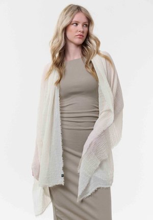 Junge Frau mit langen blonden Haaren, die ein eng anliegendes taupefarbenes Kleid und einen lockeren, strukturierten, off-white Schal trägt, schaut seitlich vor einem schlichten Hintergrund.