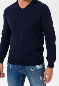 Jersey azul marino de punto con cuello en V, mangas largas y dobladillo acanalado. Presenta un pequeño logo en el pecho; combinado con jeans vaqueros desgastados.