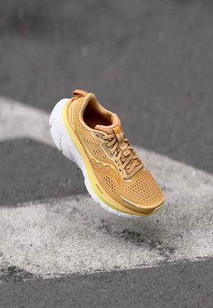 Chaussure de course marron clair avec semelle blanche suspendue au-dessus d'un bitume avec une peinture routière blanche, montrant la tige en maille et les lacets.