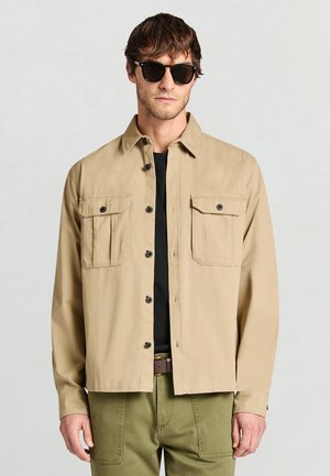 Beige button-up shirt with two front pockets, a collar, and long sleeves; worn over a black shirt with olive green pants.