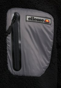 Chaqueta de forro polar negra con un bolsillo de nailon gris con cremallera. Incluye un logotipo rectangular de la marca con acentos de color.