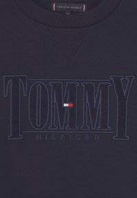 Marinblå sweatshirt med ribbad rund hals, med präglad "TOMMY HILFIGER"-logotyp över bröstet och liten röd, vit och blå rektangulär detalj.