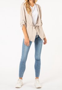 Cardigan beige à ouverture sur le devant, taille ceinturée et manches retroussées, porté sur un t-shirt blanc. Associé à un jean bleu clair et des baskets blanches.
