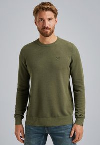 Maglione lavorato a maglia verde oliva con maniche lunghe, polsini e orlo a coste, motivo testurizzato e piccolo logo sul petto. Indossato con jeans blu.