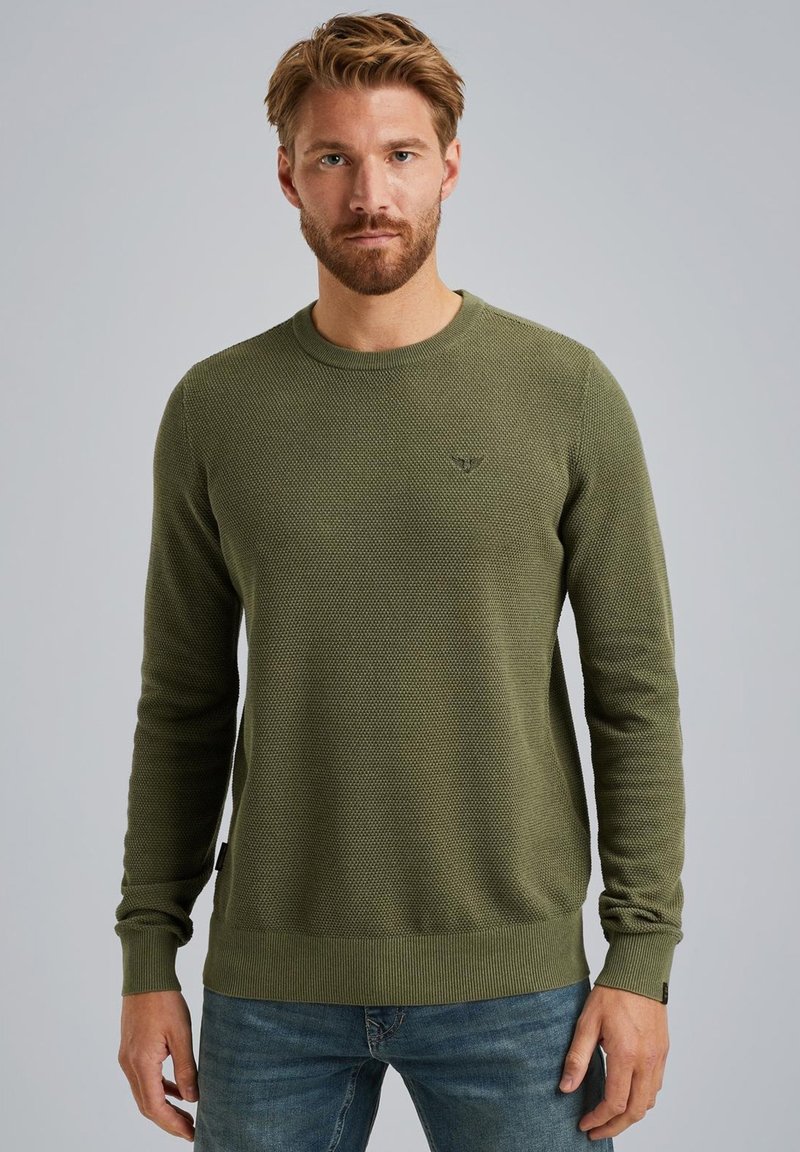 Maglione lavorato a maglia verde oliva con maniche lunghe, polsini e orlo a coste, motivo testurizzato e piccolo logo sul petto. Indossato con jeans blu.