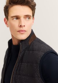 Gestepptes Weste mit einem dunklen, gemusterten Äußeren, braunen Lederakzenten auf den Schultern und einem hohen Kragen. Trägt es über einem marineblauen Pullover.