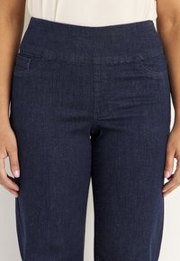 Nainen, jolla on ylhäiset tummansiniset farkut etutaskuilla ja valkoinen paita, kädet rentoina sivuilla.