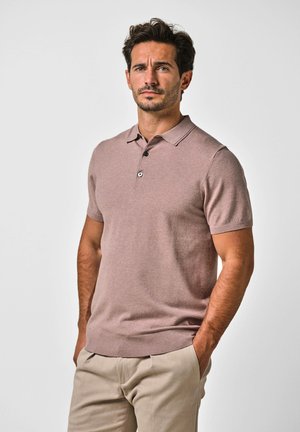 Mann mit kurzem dunklem Haar und leichtem Bart, der ein kurzärmeliges Poloshirt in Mauve und beige Hose trägt, steht mit den Händen in den Taschen.