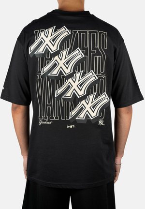 Person trägt ein schwarzes T-Shirt mit großen, sich überlappenden weißen New York Yankees-Logos und stilisiertem "YANKEES" Text auf der Rückseite.