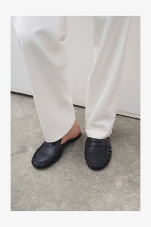 Inimene, kes kannab valgeid pükse ja musti nahast slip-on mokasiine, seisab betoonpõrandal.