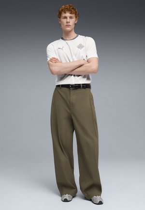 Joven con cabello rizado de pie con los brazos cruzados, vistiendo una camiseta blanca de Puma, pantalones anchos de pierna en color oliva, cinturón negro y zapatillas grises.