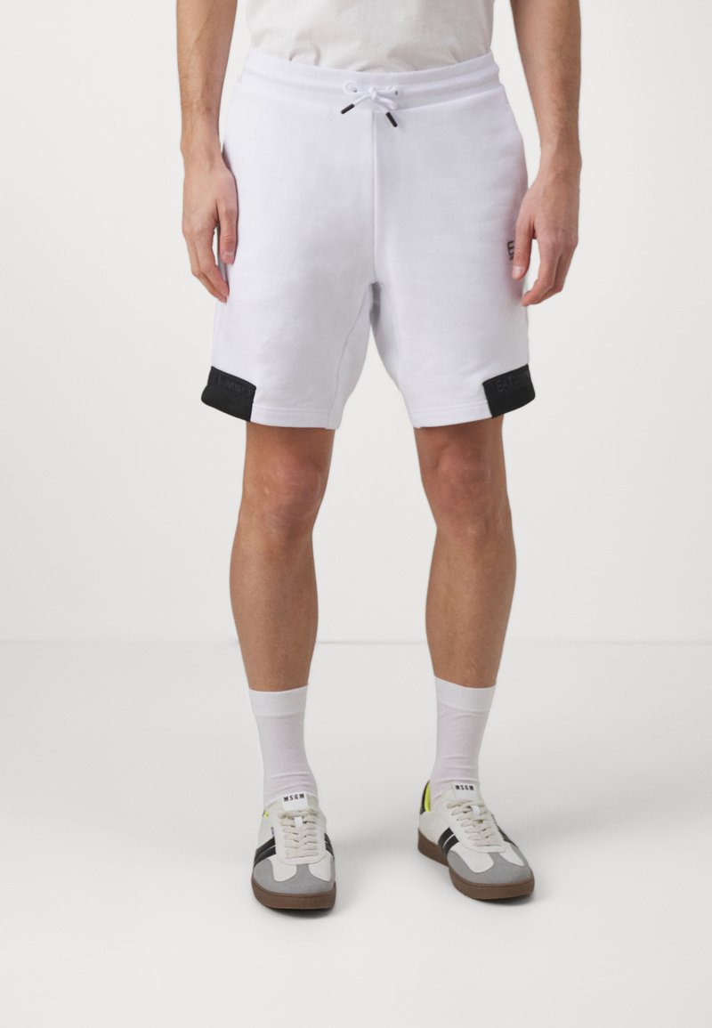 Pantalones cortos de algodón blancos con acentos negros en el dobladillo, cintura elástica con cordón y un ajuste relajado. Llevados con zapatillas grises y blancas.