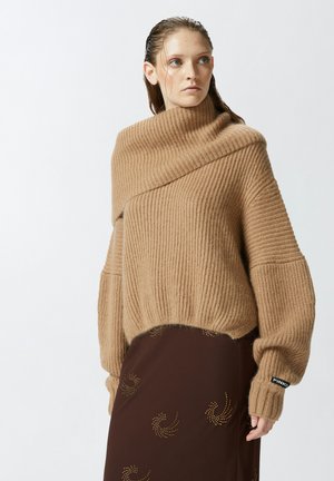 Maglione beige oversize a coste con colletto alto, maniche ampie e finitura testurizzata. Accentuato da un'etichetta con logo nera sul polsino.