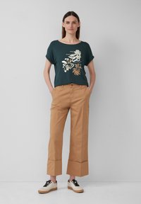Donkergroene shirt met korte mouwen en metallic bloemenprint, gecombineerd met tan brede pantalon en beige sneakers met zwarte veters.