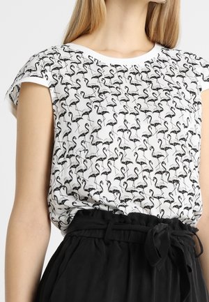 Haut blanc à manches courtes avec motif flamant noir porté rentré dans un pantalon noir taille haute style paperbag avec ceinture nouée.