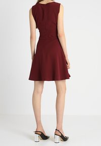 Bordeaux-farbenes, ärmelloses Kleid mit tailliertem Oberteil und schwingendem Rock, ausgestattet mit einem diskreten Reißverschluss auf der Rückseite und glatter Stofftextur. Schwarze High Heels mit klobigen, metallischen Absätzen.
