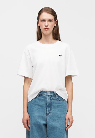 Λευκό βαμβακερό t-shirt με στρογγυλή λαιμόκοψη και κοντά μανίκια, με μικρή μαύρη ετικέτα μάρκας στο στήθος. Φορεμένο με ανοιχτό μπλε τζιν παντελόνι.