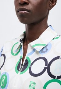 Gros plan sur une personne portant une chemise à col avec une fermeture éclair, ornée de motifs circulaires verts, bleus et noirs sur un fond blanc.