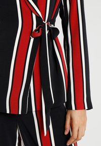 Blouse rayée rouge, noire et blanche avec manches longues et détail de taille nouée. Tissu lisse avec une texture légère et un design structuré.