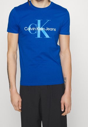 Blauwe katoenen t-shirt met korte mouwen, featuring een groot lichtblauw "CK" logo en de tekst "Calvin Klein Jeans" gedrukt aan de voorkant.