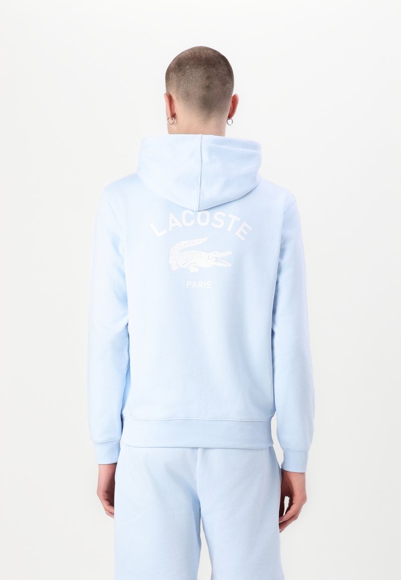 Personne portant un sweat à capuche bleu clair et un pantalon de jogging, vue de dos, avec le logo et le texte Lacoste blanc au dos du sweat.