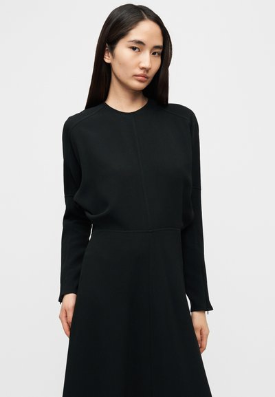 Μαύρο φόρεμα μακρυμάνικο με στρογγυλή λαιμόκοψη και εφαρμοστή γραμμή στο μπούστο, στενεύοντας σε φούστα σε σχήμα Α. Ομαλή, ματ υφή και μινιμαλιστικός σχεδιασμός.