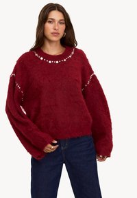 Pull rouge en peluche à coupe décontractée, avec des clous ronds le long du col et des manches. Associé à un jean en denim noir.