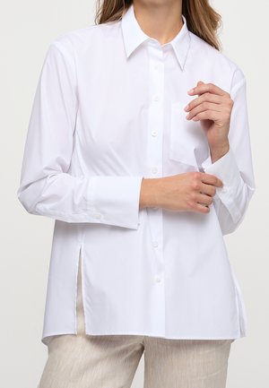 Donna che sistema il polsino di una camicia bianca con collo e bottoni, indossata con pantaloni beige chiaro su uno sfondo neutro.