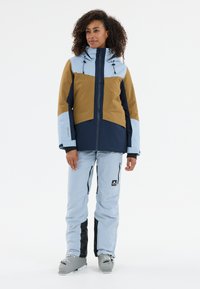 Skidjacka med bruna, marinblå och ljusblå paneler; har huva, dragkedjor och justerbara snoddar. Ljusblå skidbyxor med matchande design.