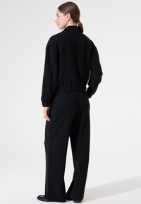 Zwarte trainingsoutfit met een slim fit jas en wijde broekspijpen. Zacht materiaal, ribbelmanchetten en tailleband. Minimalistisch ontwerp zonder zichtbare patronen.