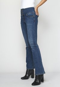 Uppflutna mörkblå denimjeans med hög midja, iögonenfallande tre dekorativa metallknappar och kontrasterande sömnad, kombinerat med svarta klackstövlar.