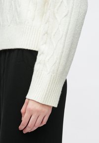 Suéter blanco de punto con puños acanalados, que muestra una textura suave. Llevado sobre tela negra, resaltando el contraste entre colores.