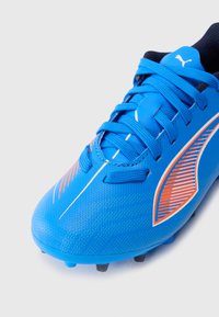 Botín de fútbol azul con parte superior sintética texturada, acentos naranjas en contraste, cordones planos y una suela exterior de goma con tacos para mayor tracción.