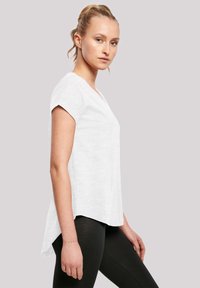 Weißes kurzärmliges V-Ausschnitt-Top mit einem etwas längeren, abgerundeten Saum hinten. Aus leichtem Stoff mit glatter Textur. Kombiniert mit schwarzen Leggings.